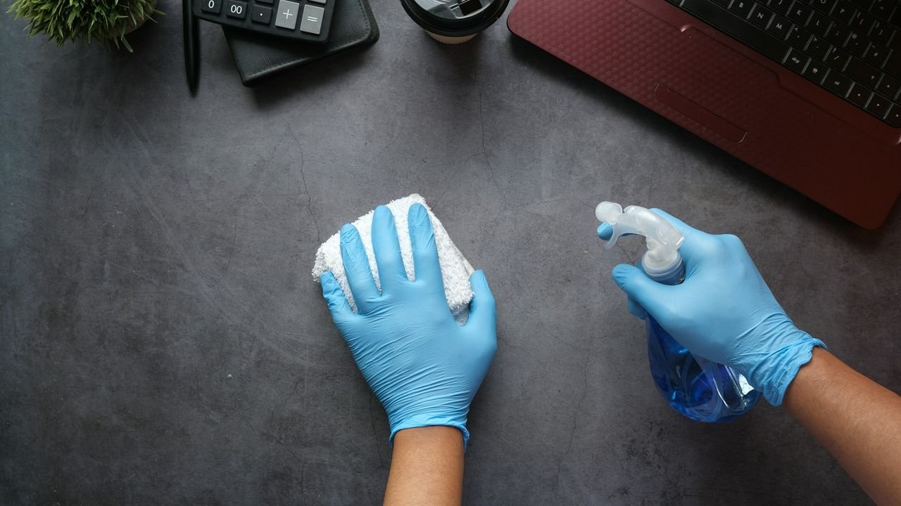 Gloved hands cleaning an office desk with a spray bottle and cloth